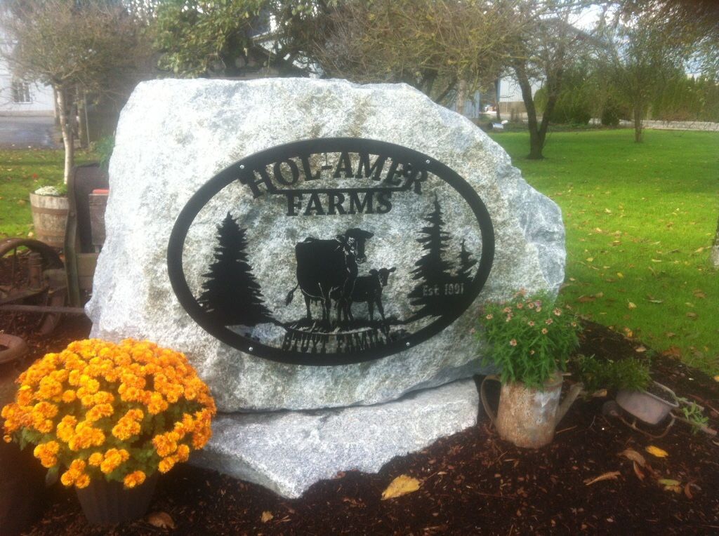 Stone farm sign with oval metal emblem, orange flowers, and green grass in a landscaped outdoor setting