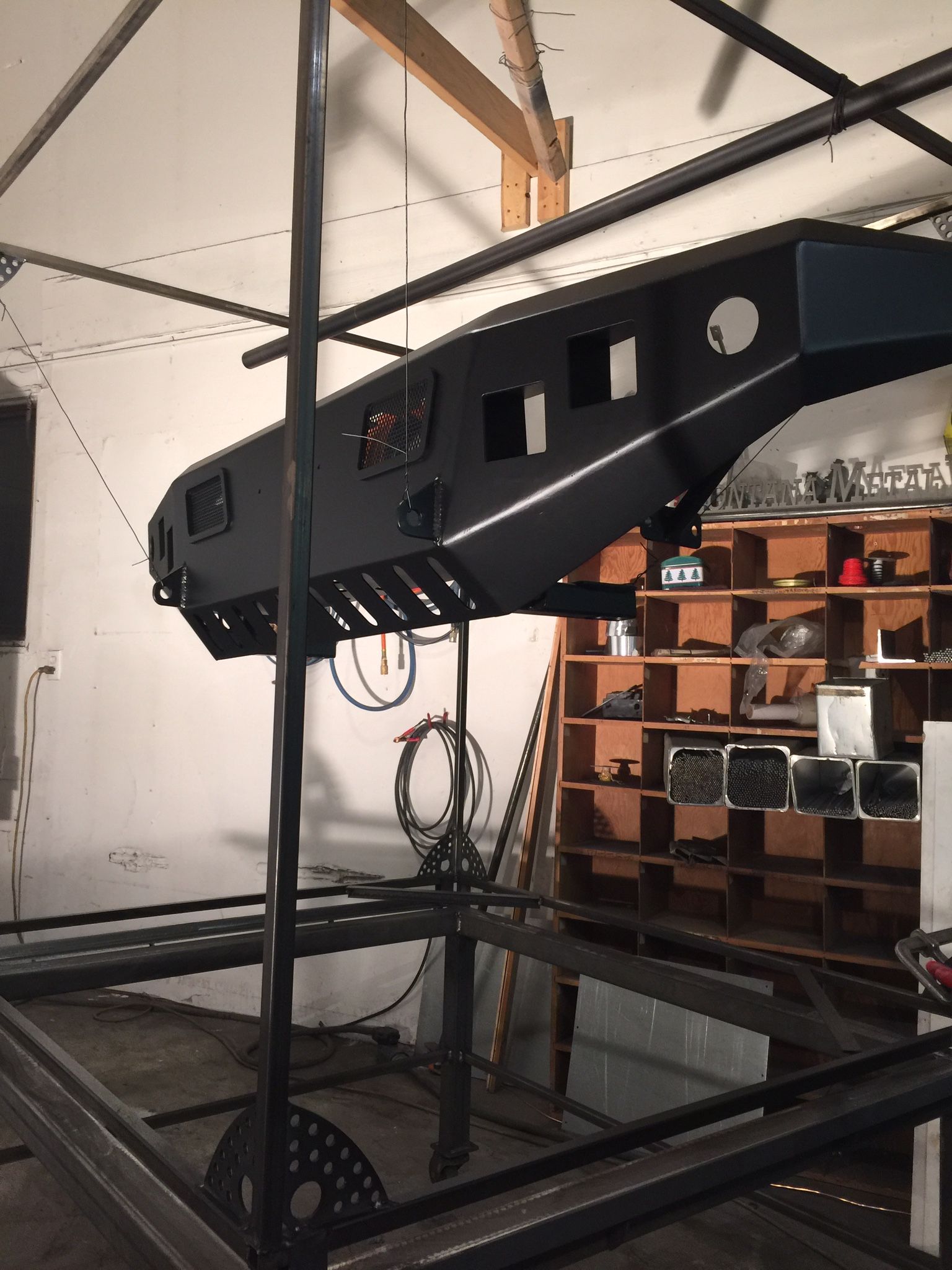 Black overhead 3D printer frame in a workshop, with shelves and storage bins in the background