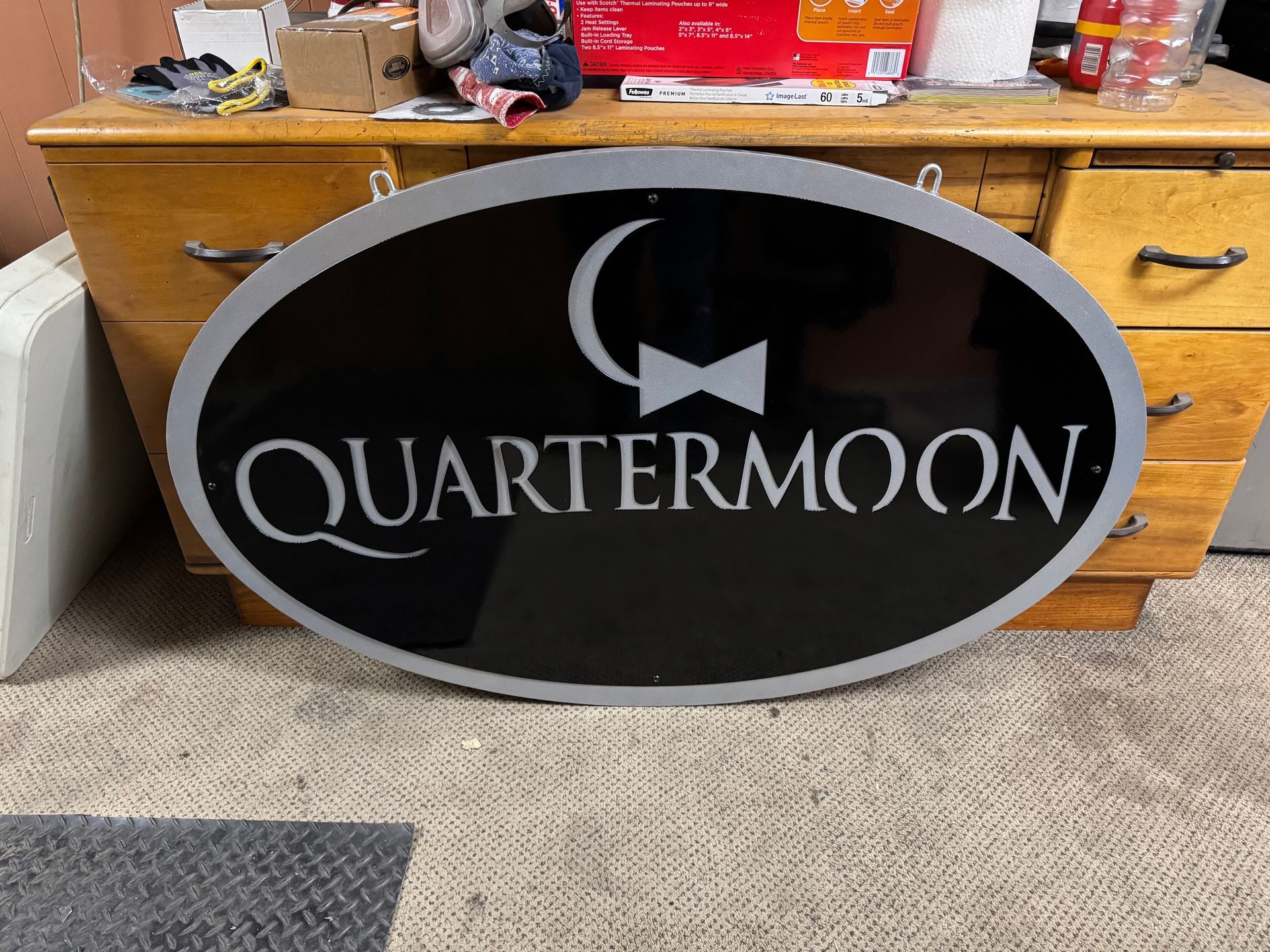 Oval black-and-silver QuarterMoon sign leaning against a wooden dresser on carpet.