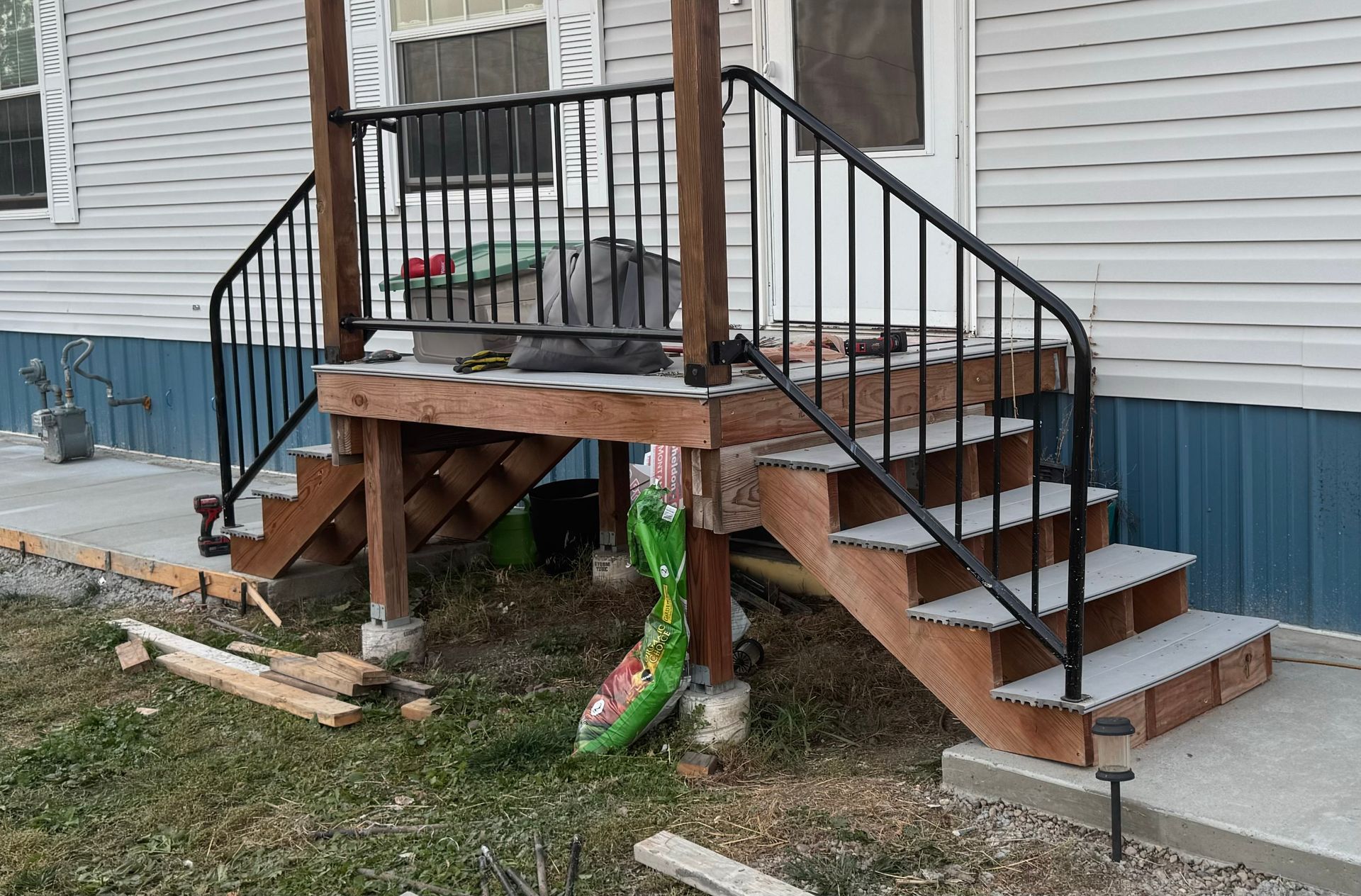 Wooden front porch stairs and deck attached to a house, with black railings and blue siding underneath.