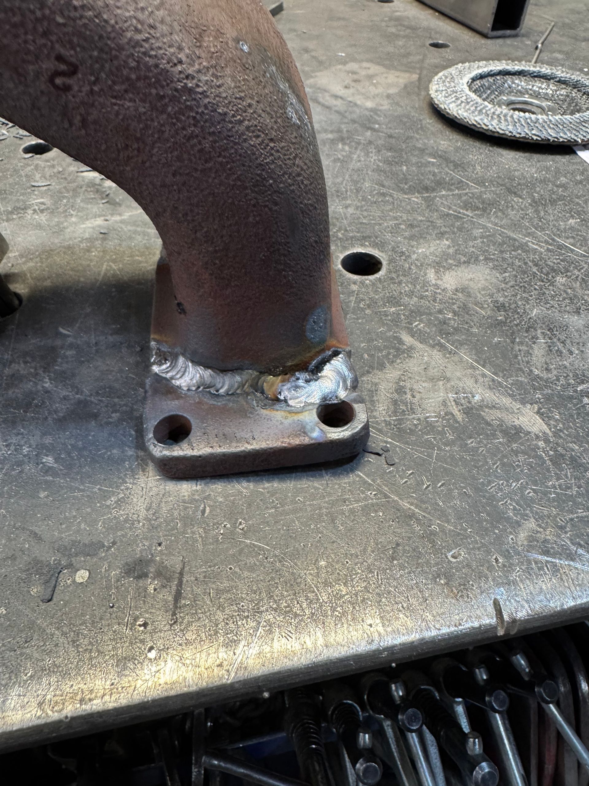Close-up of a rusted metal pipe welded to a steel plate on a machine, with bolts and metal shavings nearby