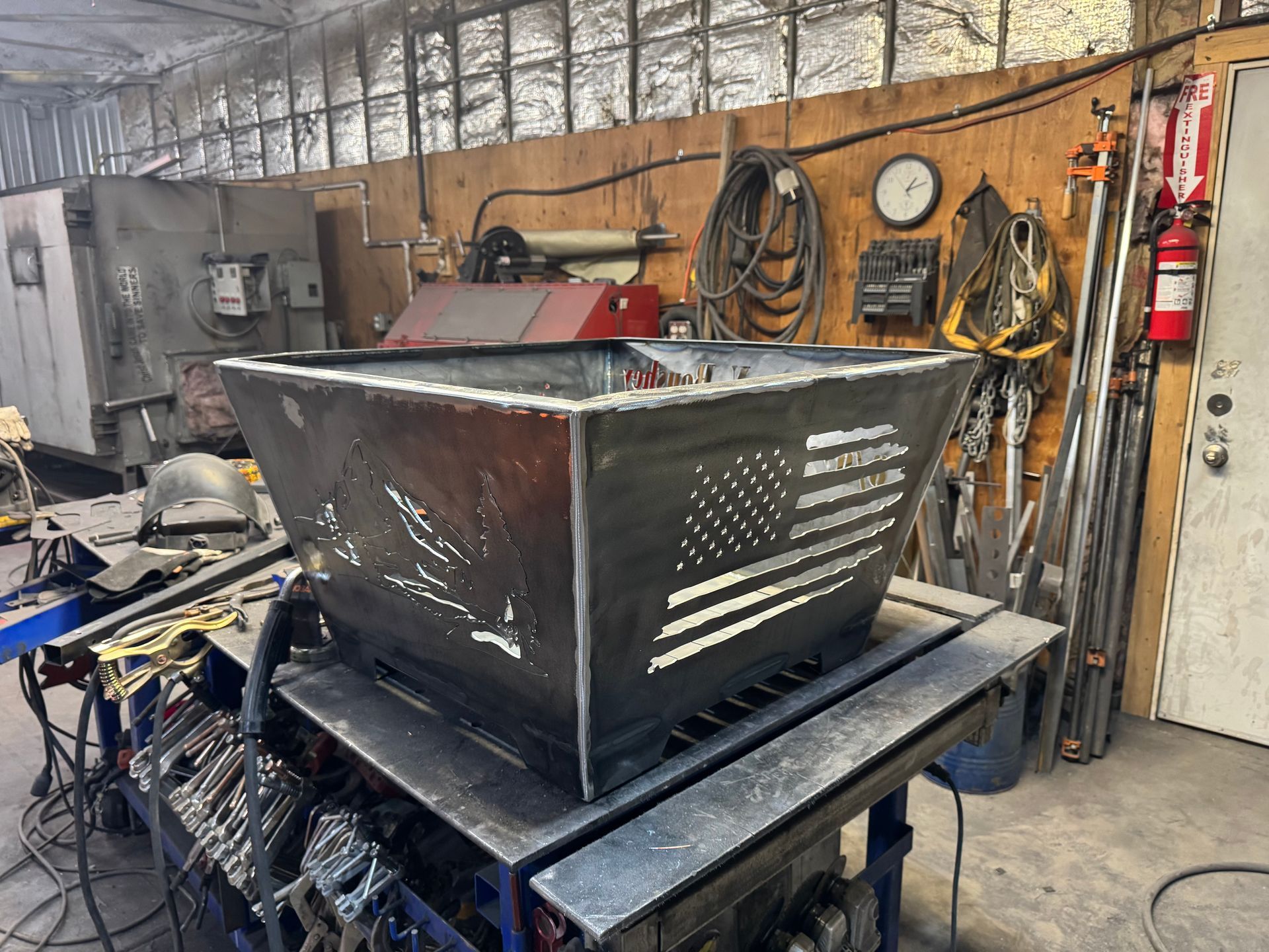 Black metal box-like machine on a worktable in a cluttered workshop with tools on the wall