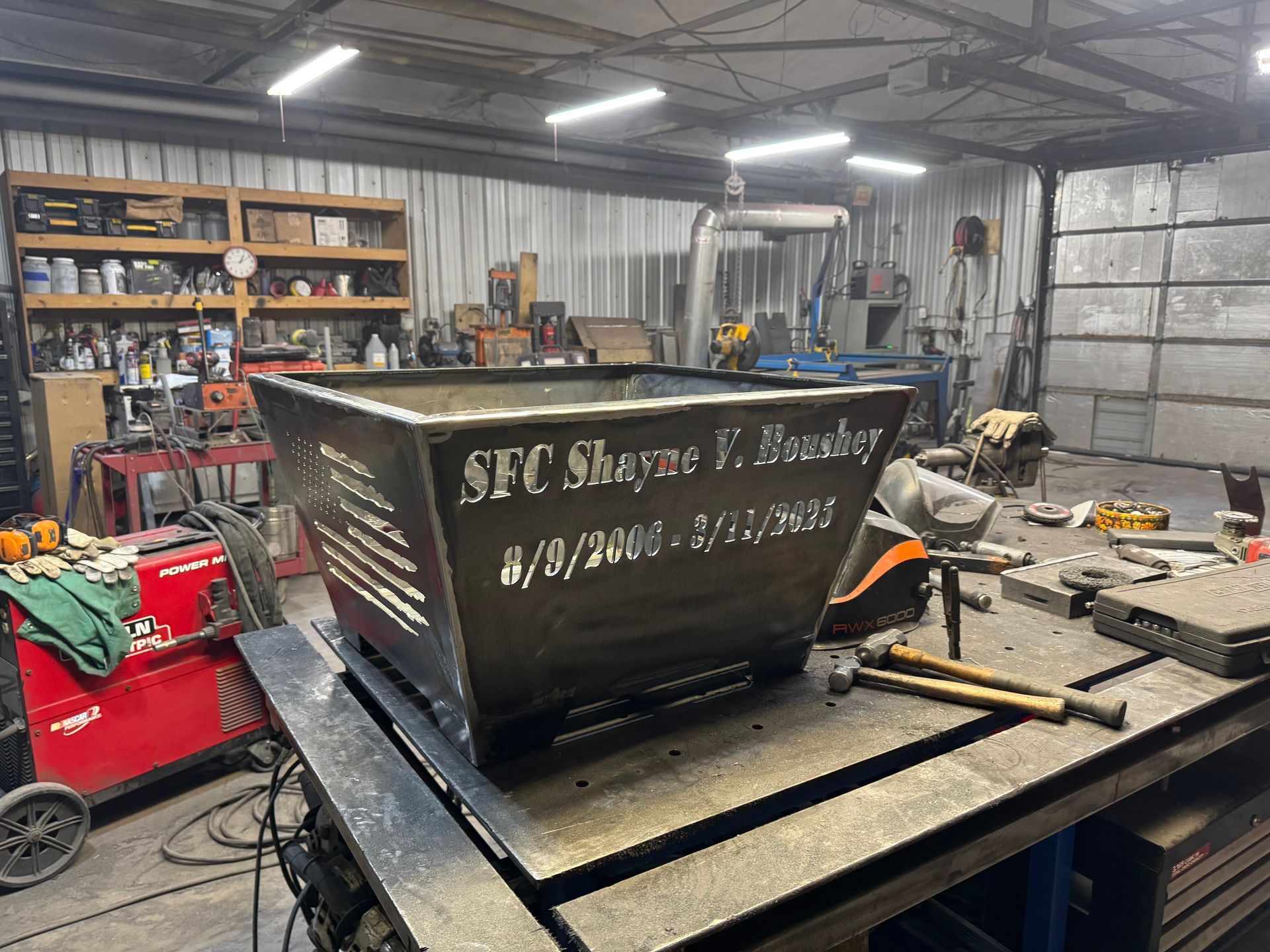 Workshop with a large black metal dumpster on a trailer, surrounded by tools and equipment.