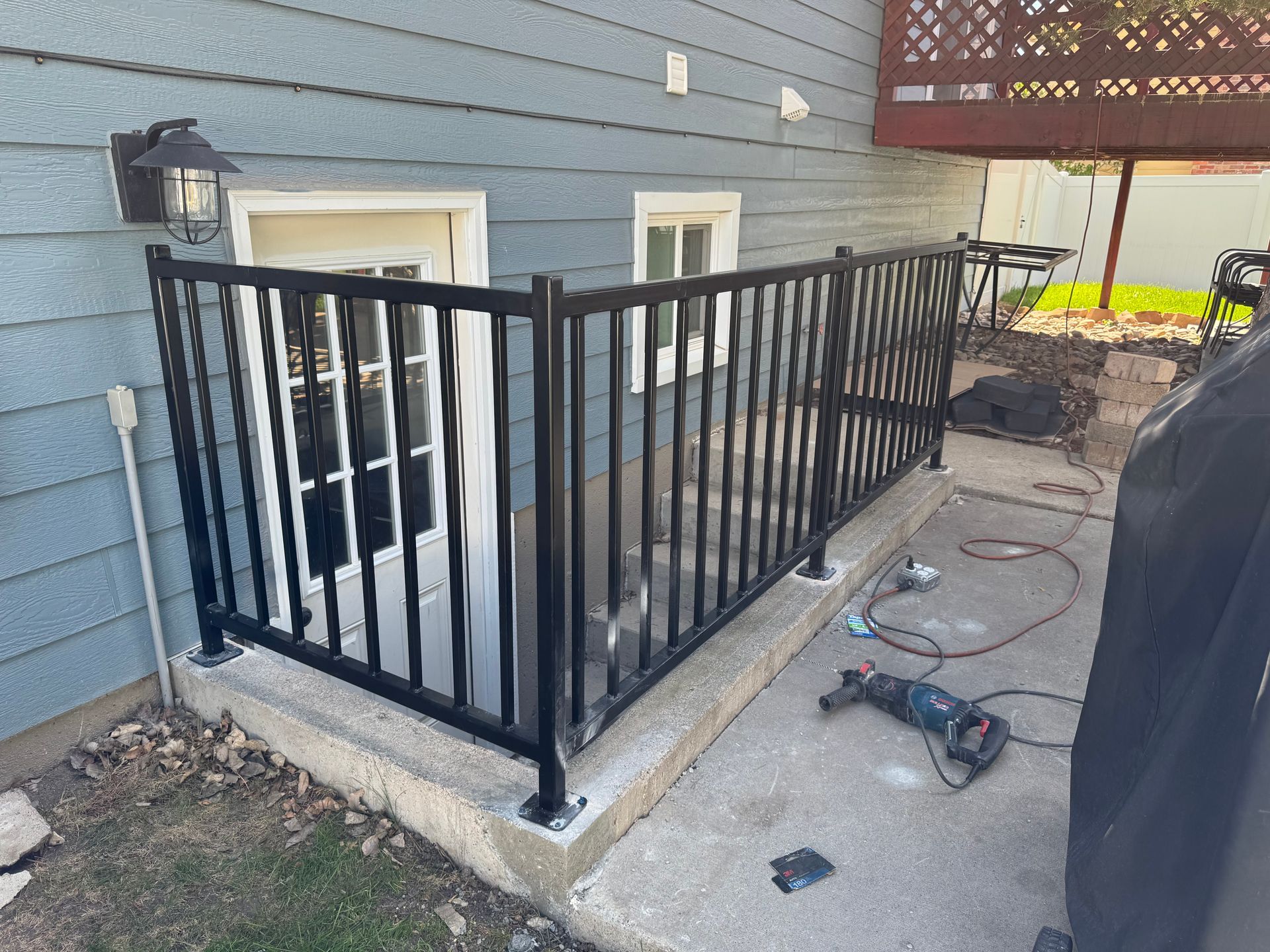 Black metal railing around a small concrete patio beside a blue house, with tools on the ground.