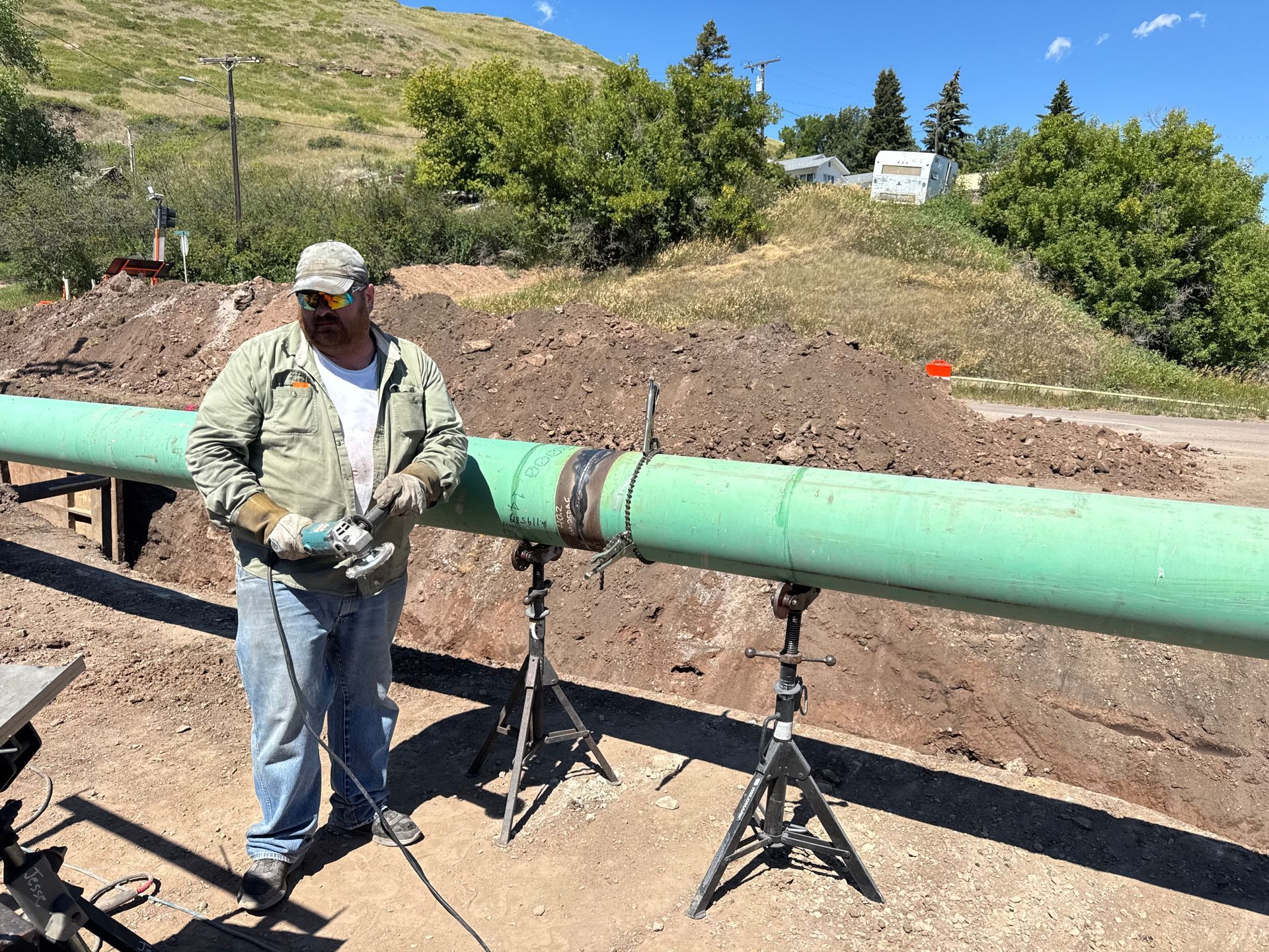 Worker standing beside a large green pipeline on supports in a dirt construction area outdoors