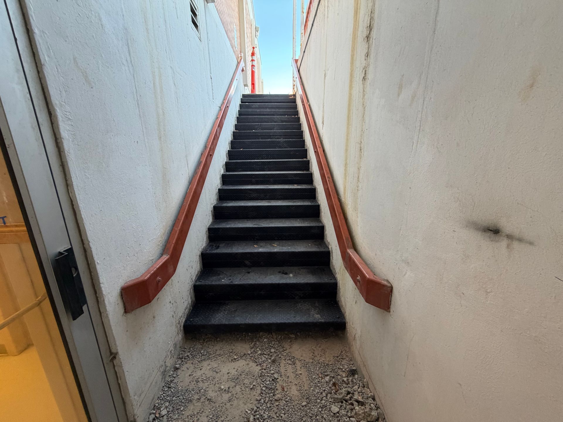 Narrow stairwell with steep black steps, red handrails, and white walls leading upward