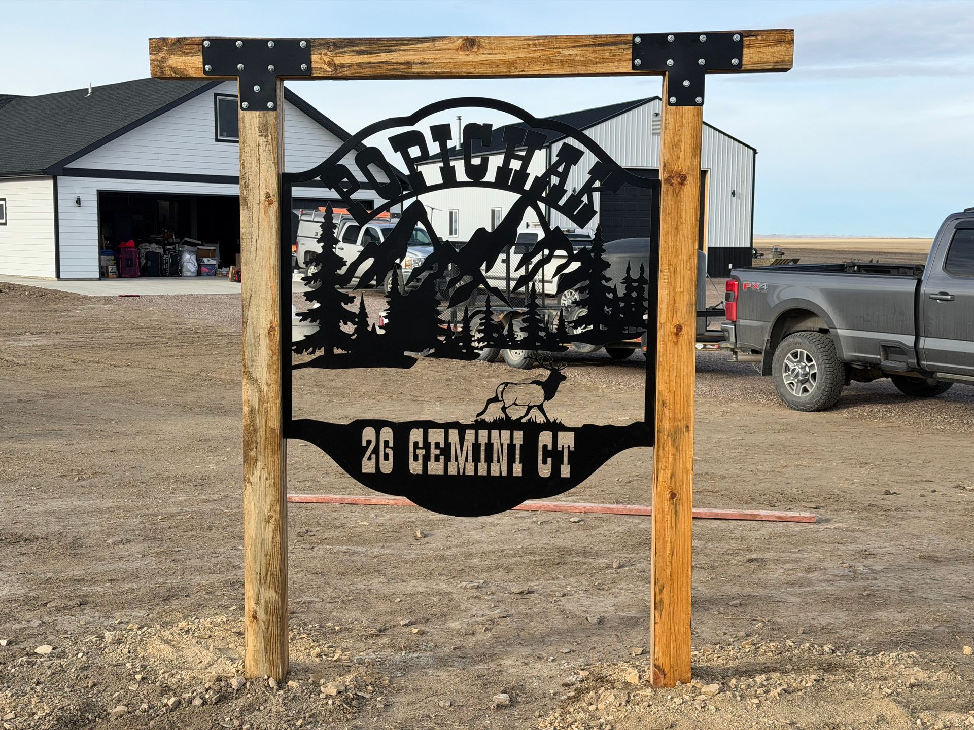 Black metal roadside sign between wooden posts in a gravel lot, with truck and barn in background.