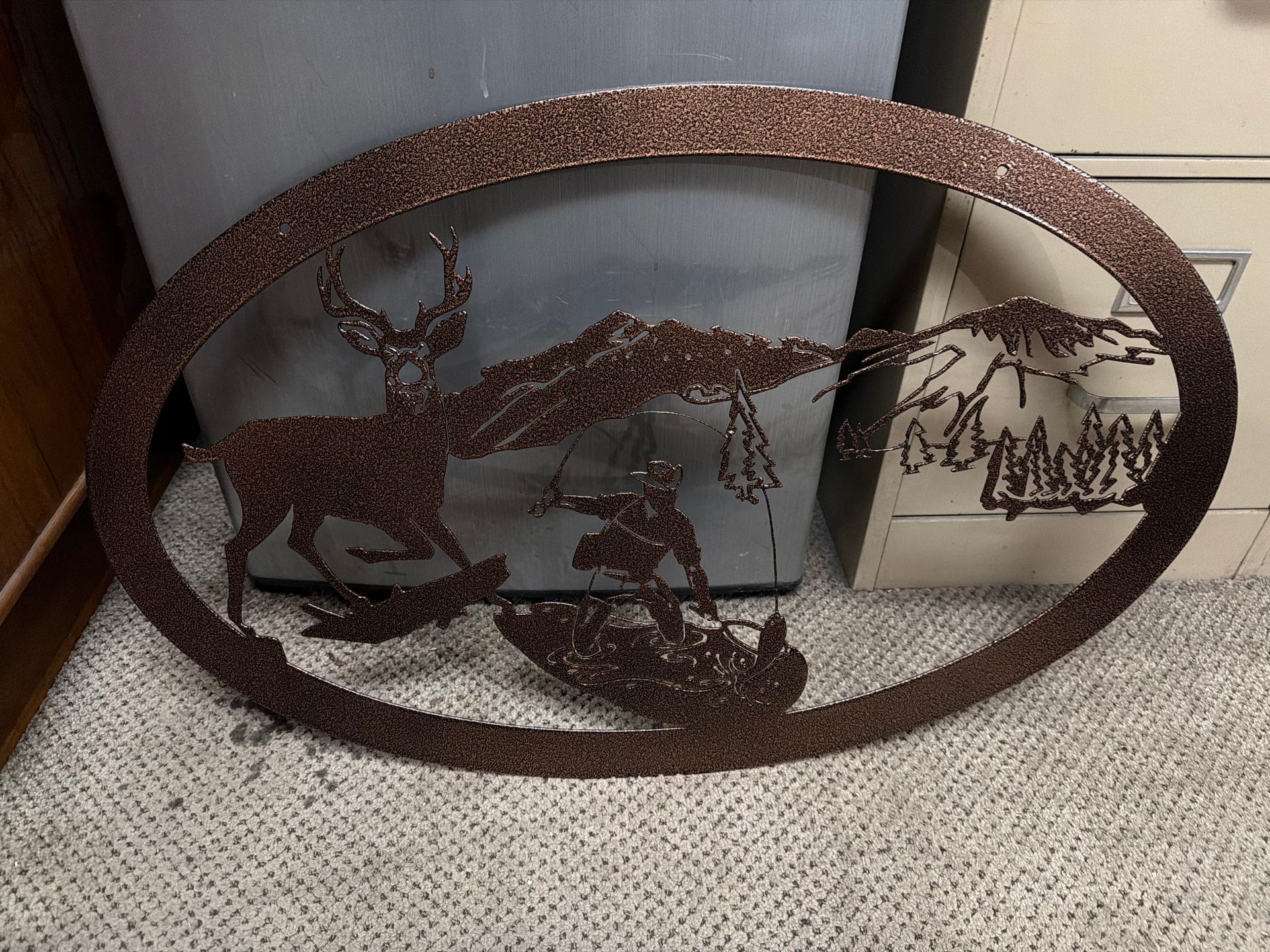 Rust-colored oval metal wall art featuring deer and forest silhouettes on a carpeted floor