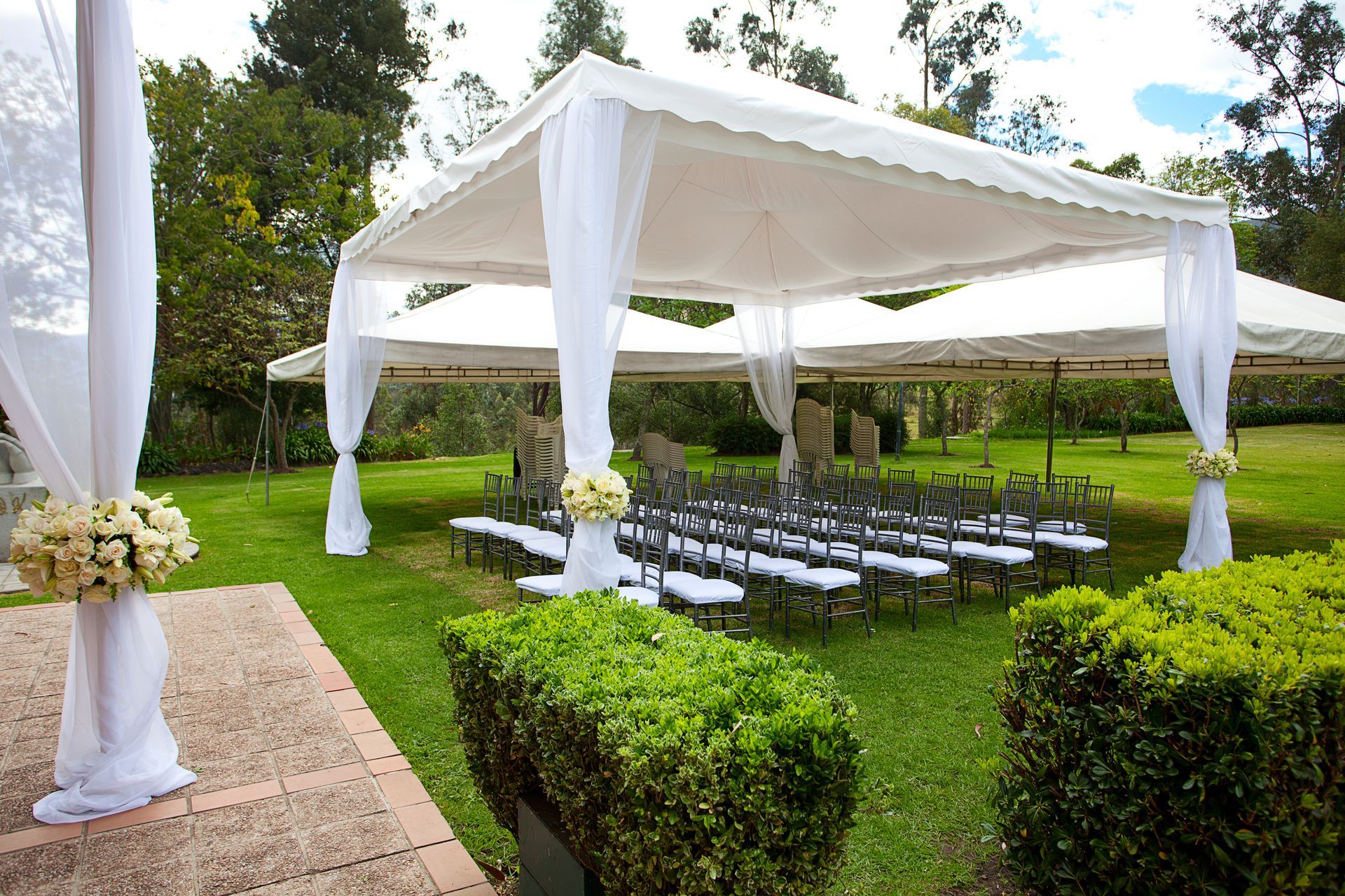 Outdoor wedding ceremony setup with white canopy, rows of chairs, and floral arrangements on a green lawn.
