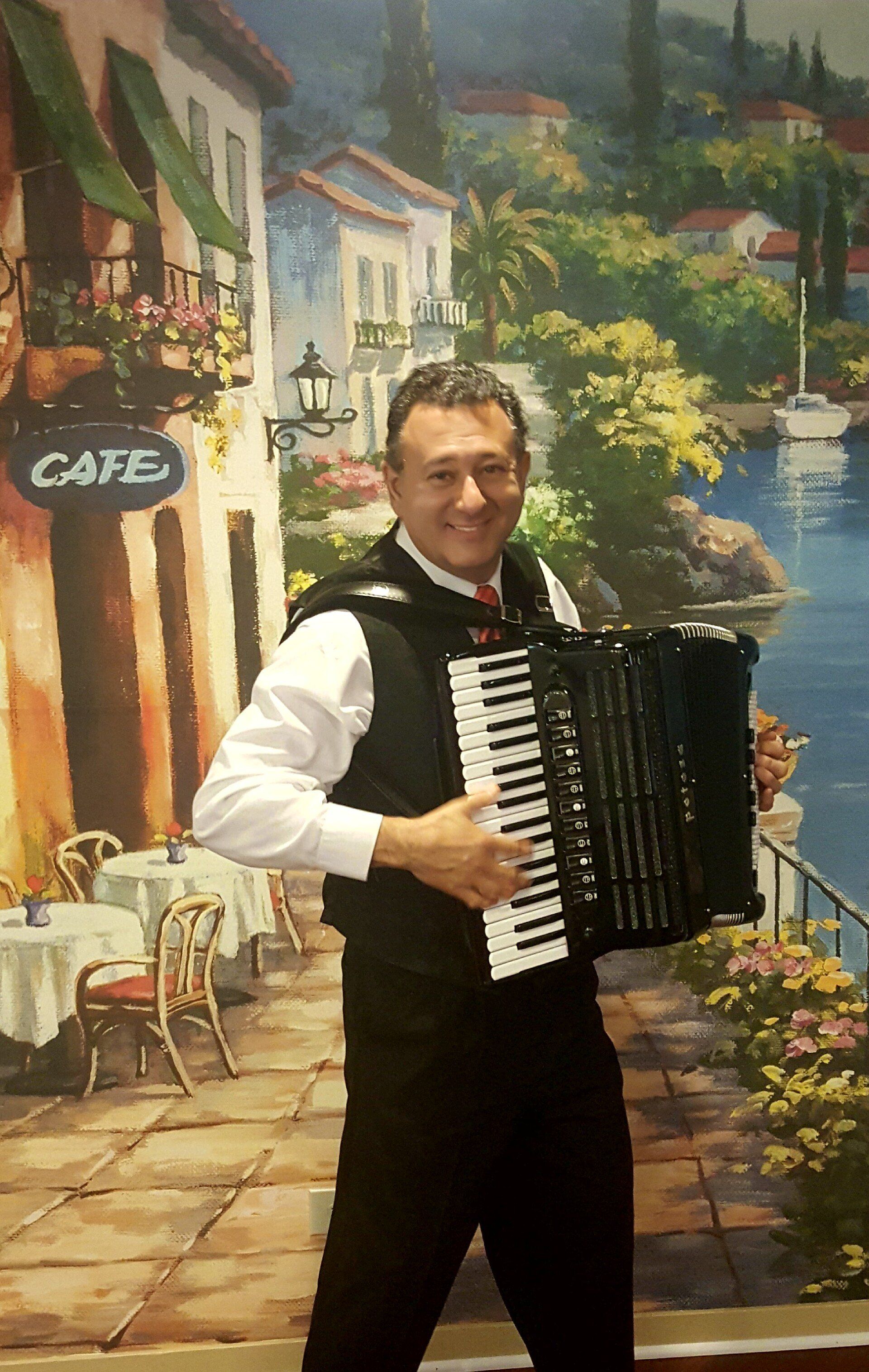 Man in vest plays accordion, smiling. Outdoors cafe scene backdrop.