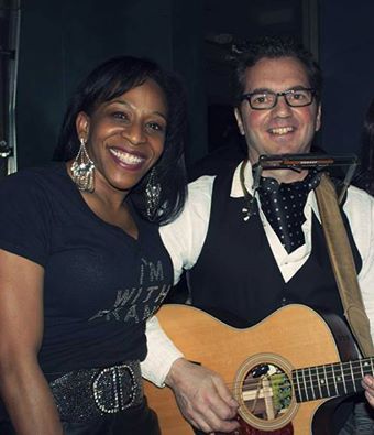 A smiling person in a black shirt poses with a musician playing acoustic guitar, inside setting.