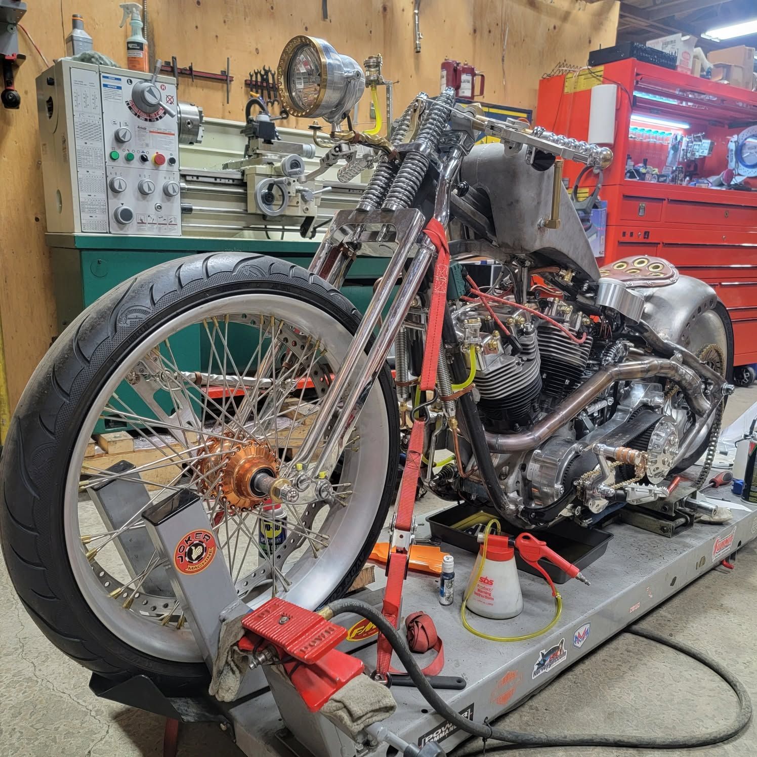 Custom chopper motorcycle on a workshop lift, with exposed frame, large front wheel, and tools around it