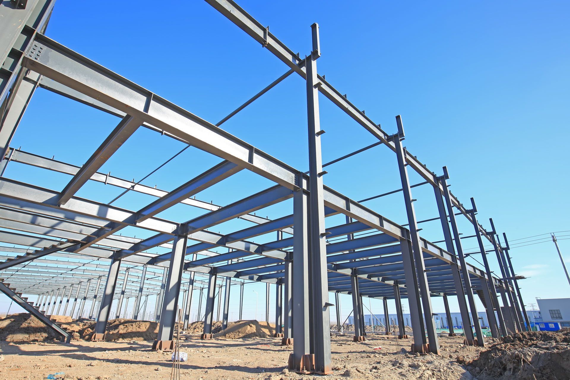 Steel building frame under construction on a sandy lot, with blue sky and scaffolding.