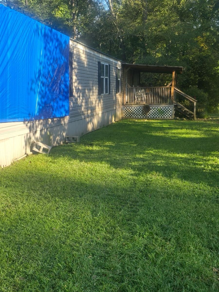 Side view of a mobile home with a blue tarp, wooden porch, and grassy yard in the sunshine.