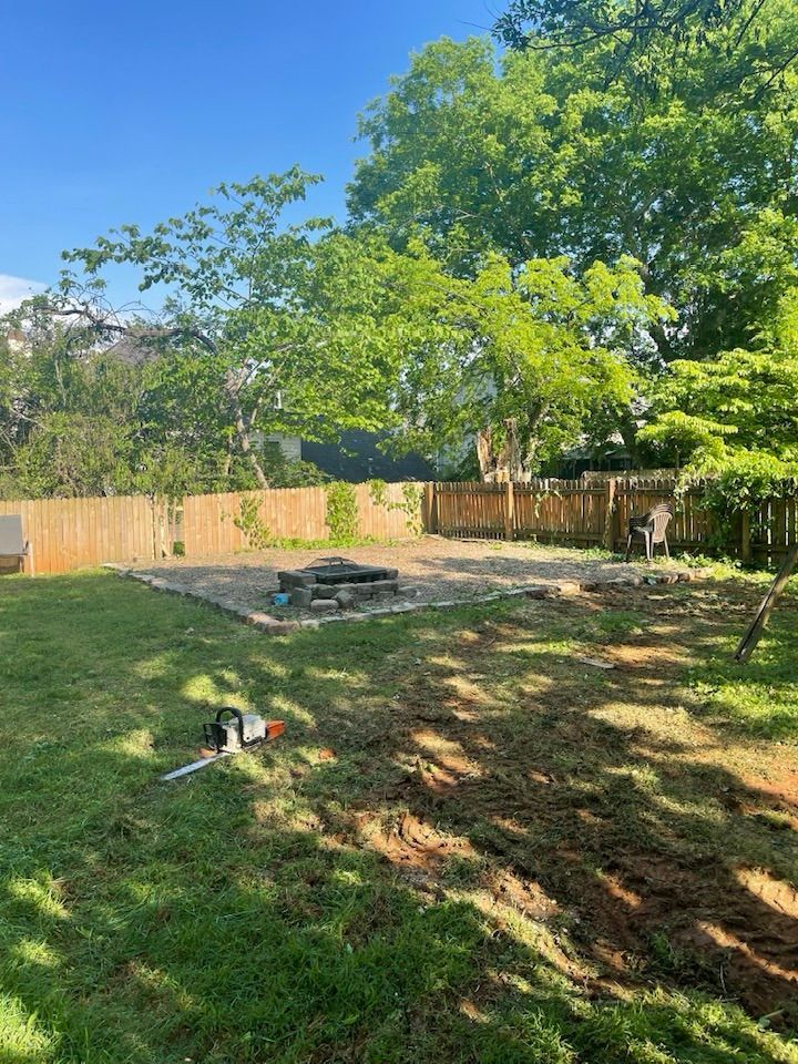 Backyard with a gravel patio and fire pit, lawn with chainsaw, and wooden fence under a blue sky.