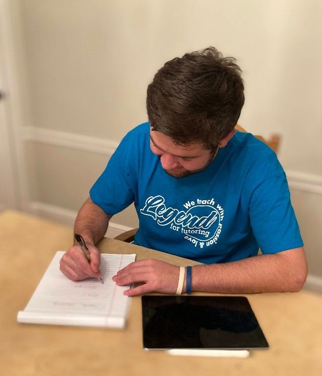 Man in blue shirt writing on paper at a table, with a tablet.