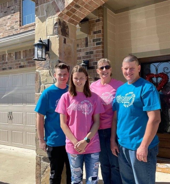 Family of four in front of a house; two in blue shirts, two in pink.