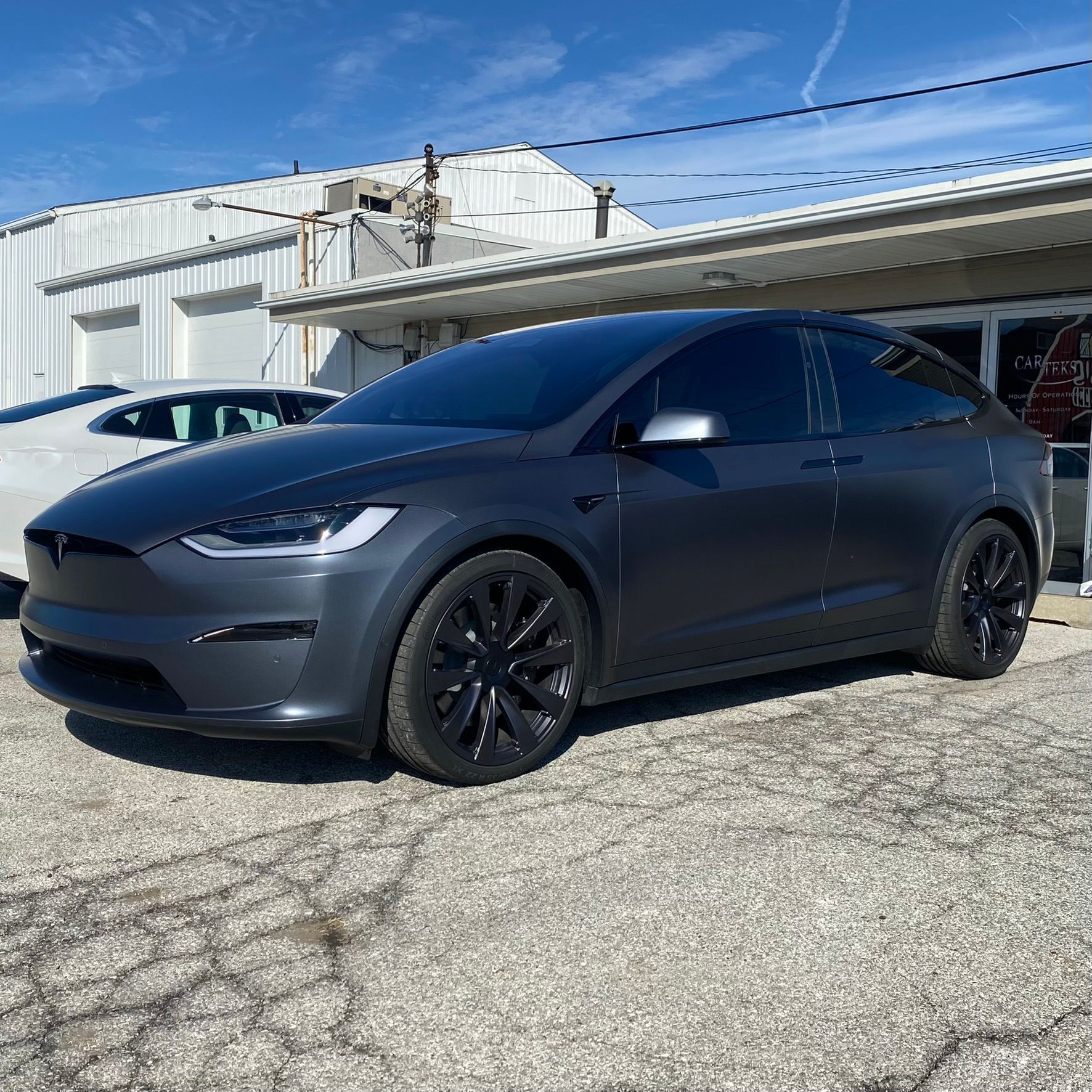 Dark gray Tesla Model X with black wheels parked in front of a building on a sunny day.