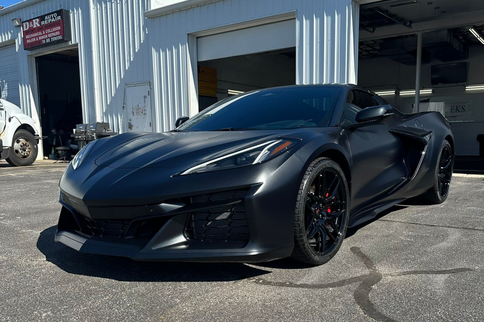 Matte black sports car parked outside a garage, bright sunlight.