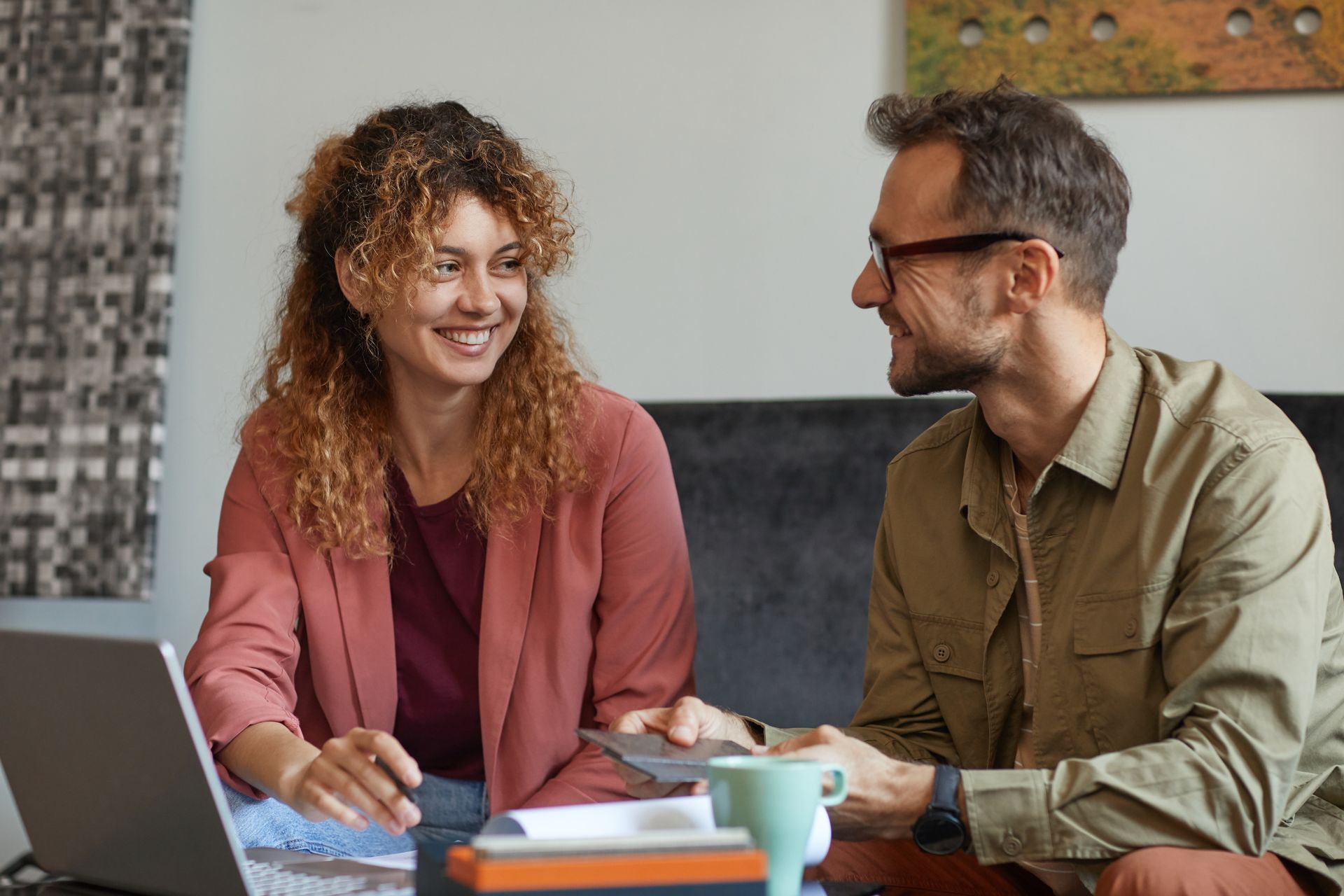 Two coworkers smiling and talking at a desk with notebooks and coffee cups