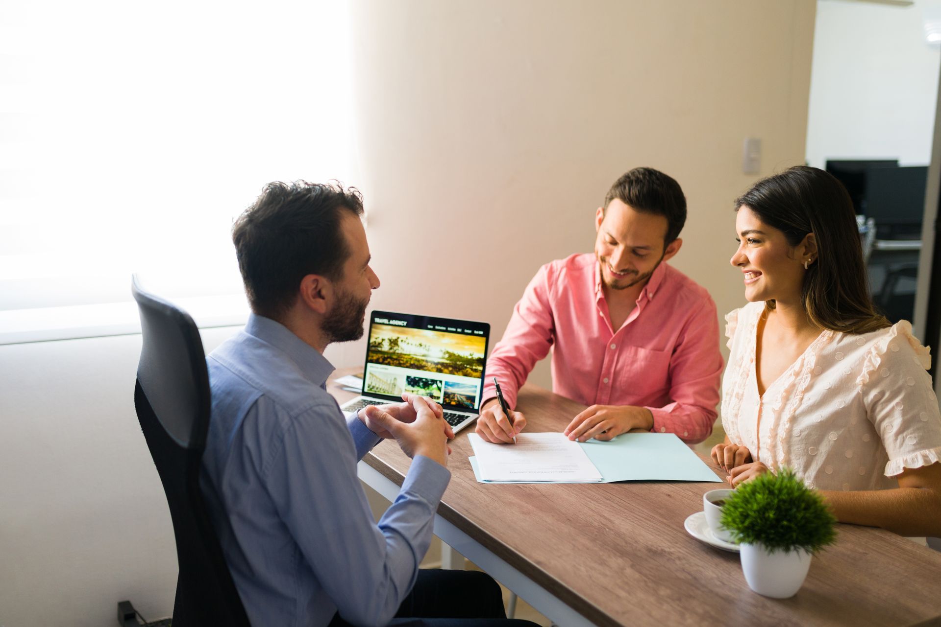 Three coworkers discuss paperwork at a wooden table, with a laptop and small plant in a bright office.