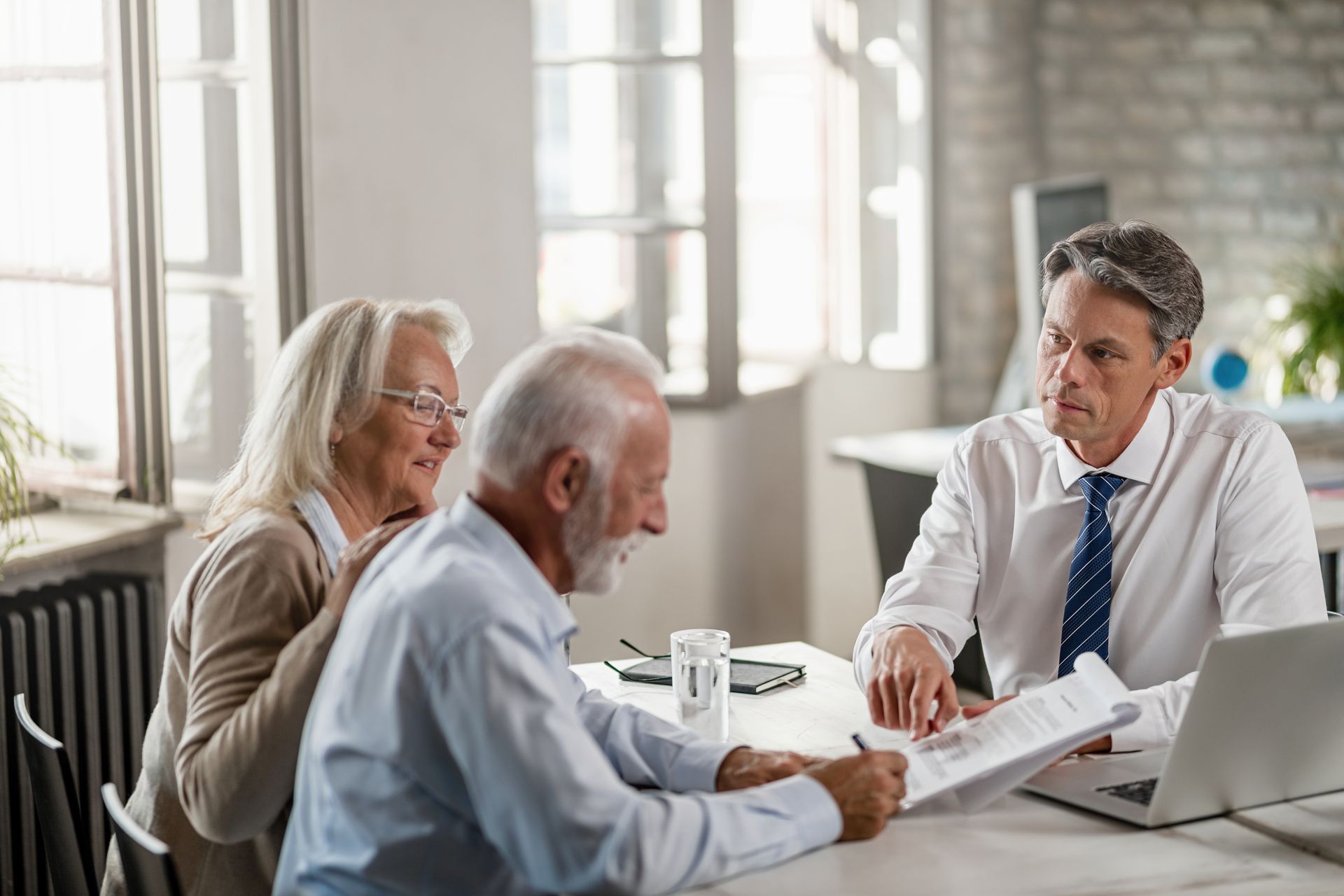 Three people in a bright office discussing documents at a table with a laptop