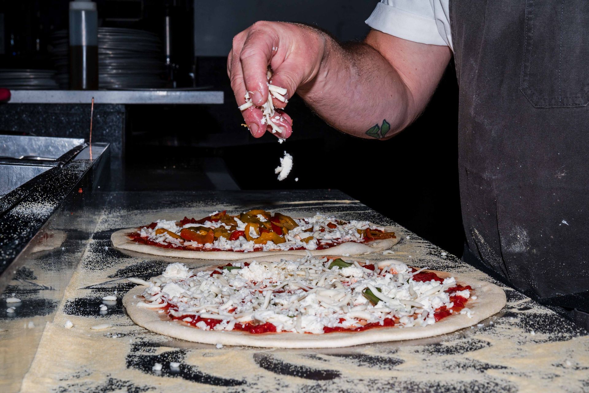 A close-up of a chef’s hand sprinkling fresh mozzarella cheese over raw pizza dough topped with tomato sauce and vegetables, capturing the handmade preparation process in a professional kitchen.