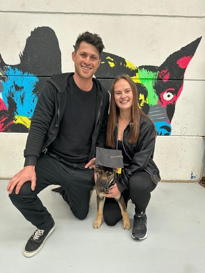 Two People Kneel With a Dog Wearing a Graduation Cap — DogEd Daycare & Training in Gosford, NSW