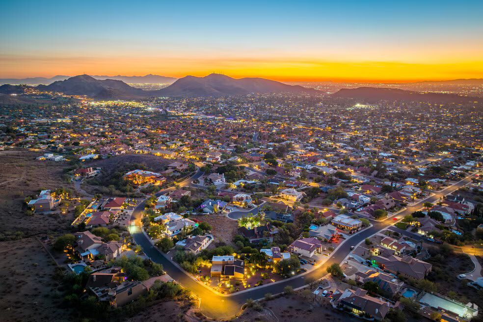 City lights at dusk, homes nestled in hilly terrain under a colorful sky.