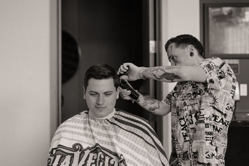 Barber cutting a man’s hair in a shop, with a striped cape and tattooed arms visible