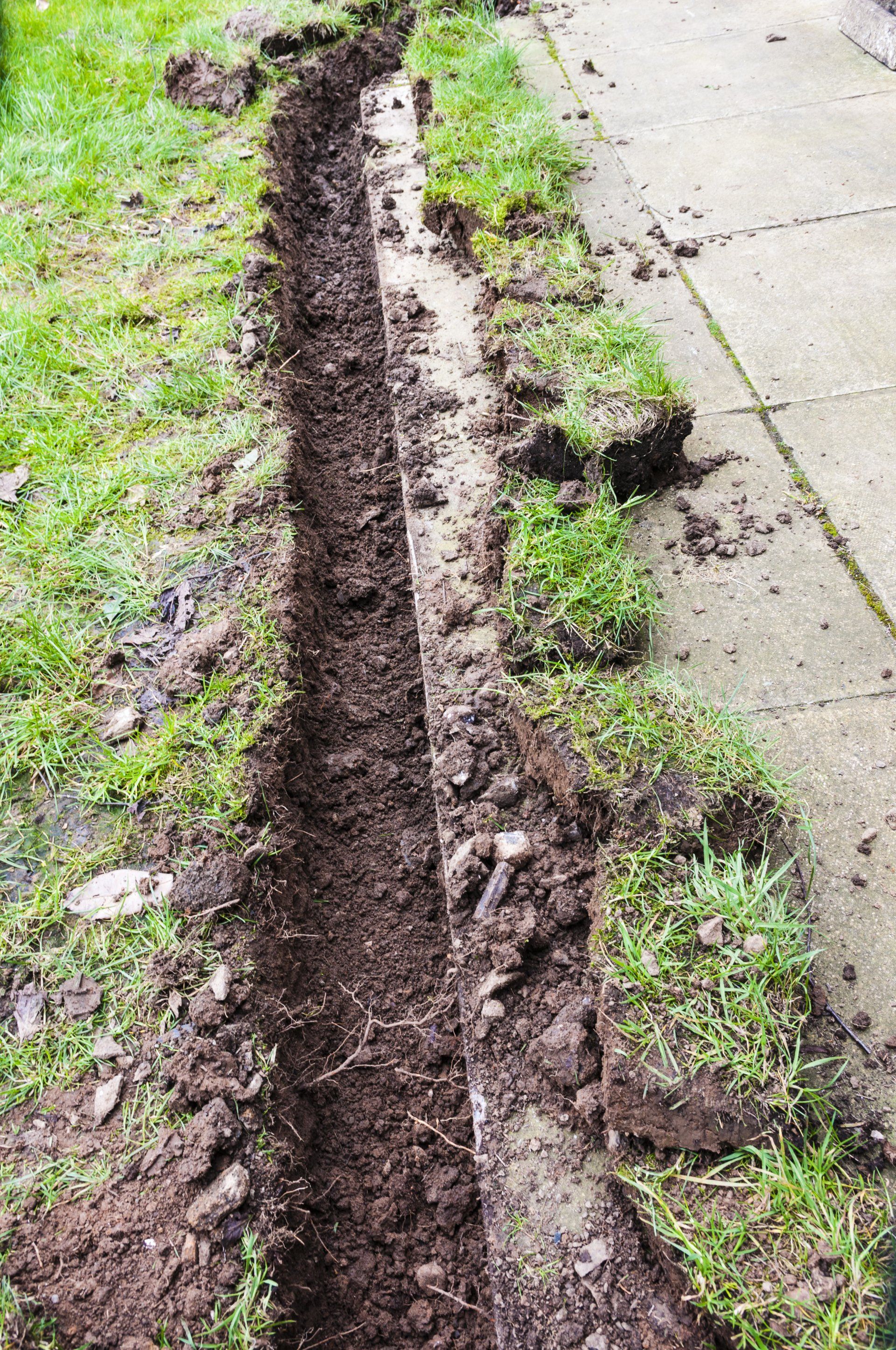 A muddy trench is being dug in the grass next to a sidewalk.