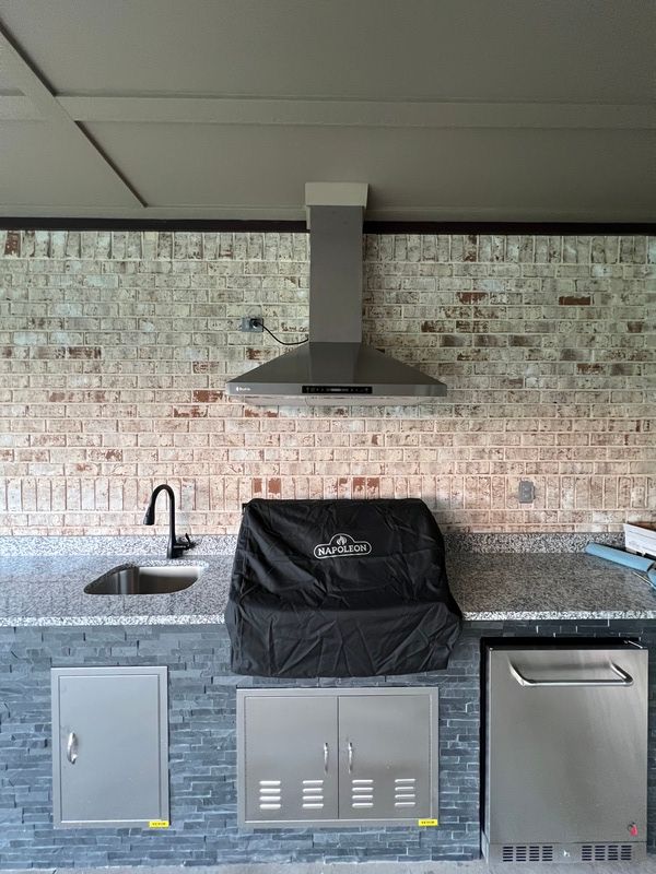Outdoor kitchen with grill under a range hood, sink, and refrigerator. Stone wall and countertop.