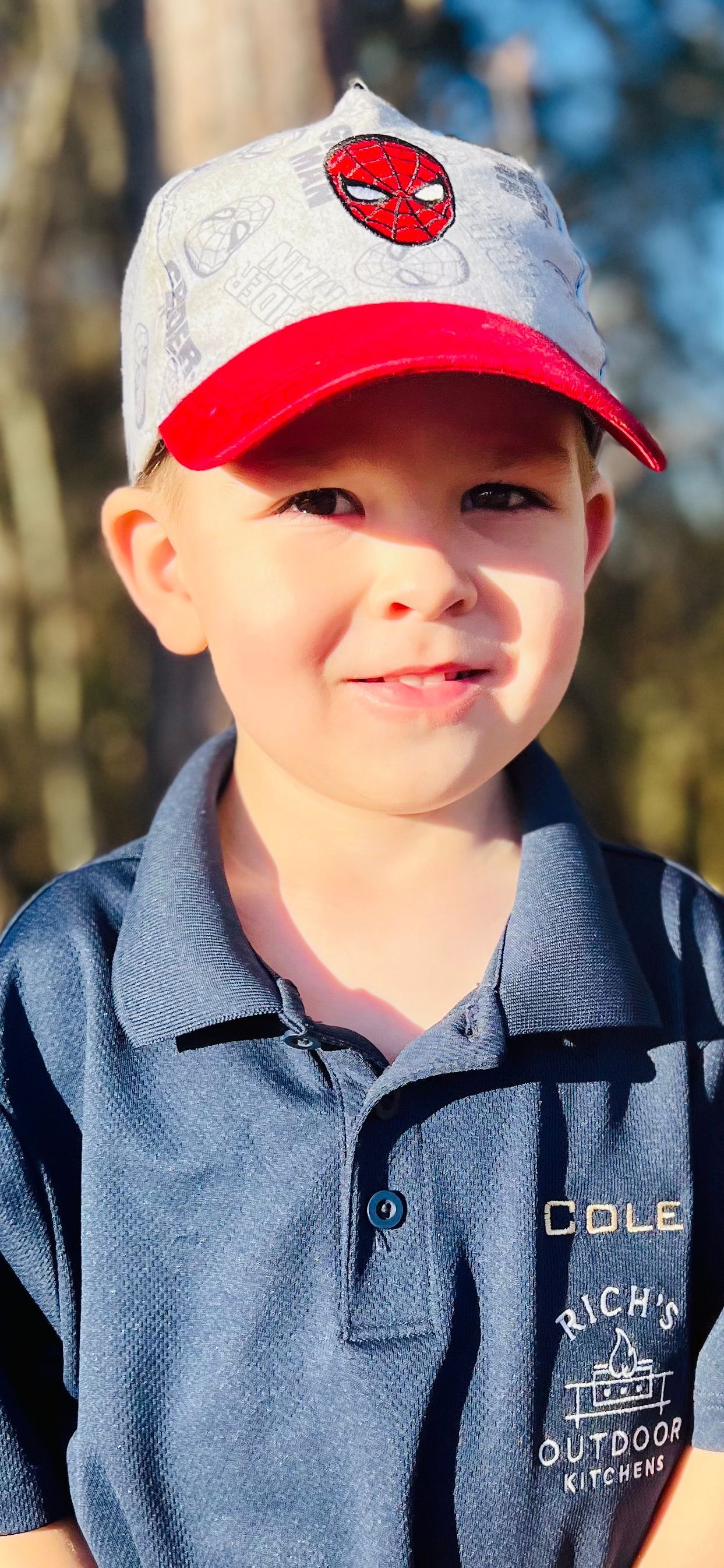 Young child wearing a Spider-Man baseball cap and a blue polo shirt, smiling outdoors.