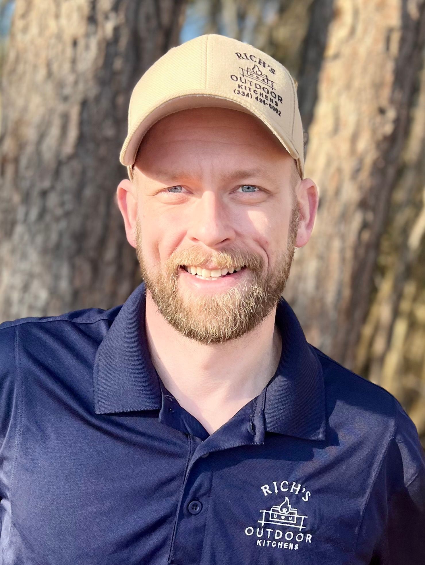 Man with a beard and a tan cap smiles. He wears a blue polo shirt, and is outdoors.
