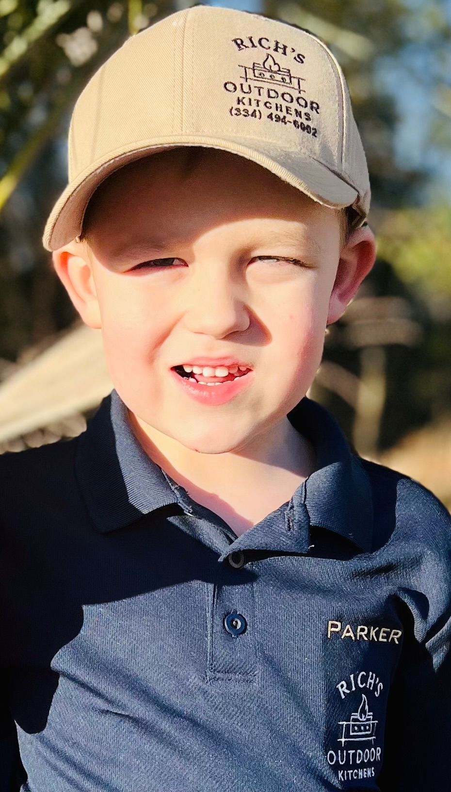 Boy in a baseball cap and polo shirt smiles outdoors.