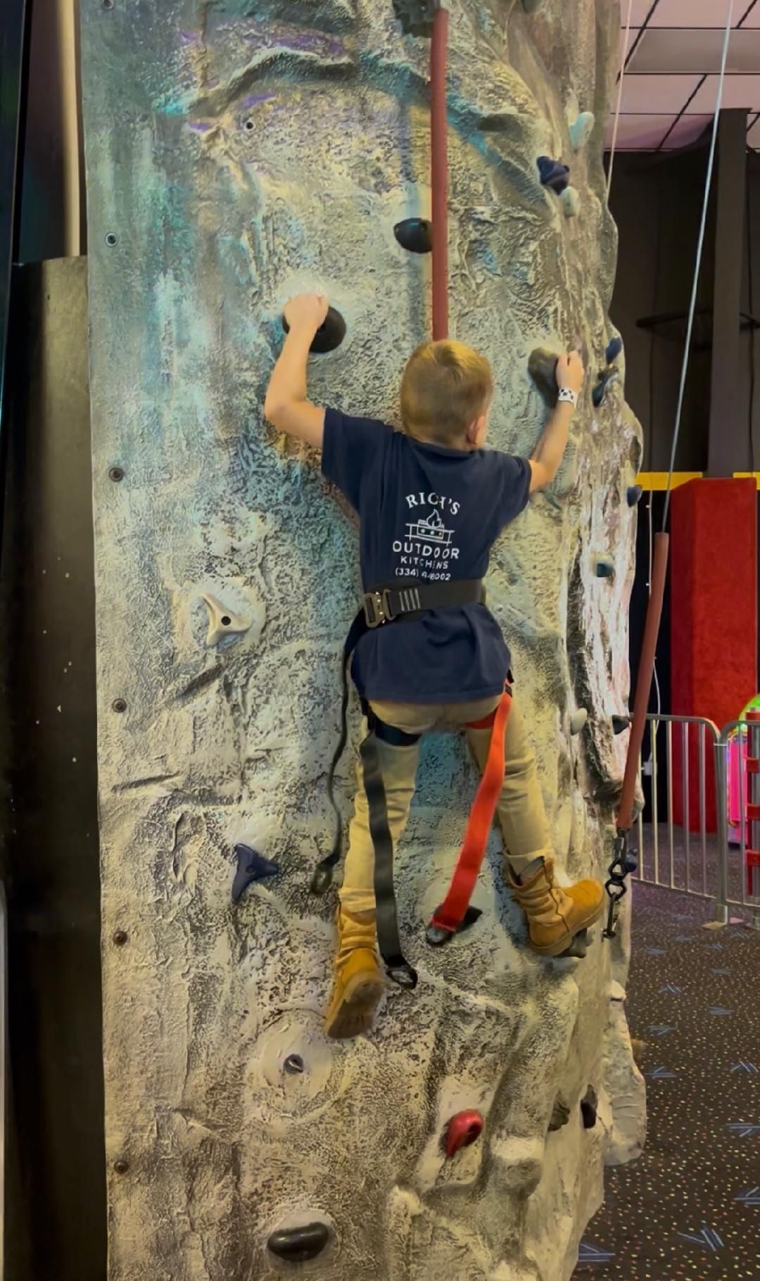 Boy in a harness climbs a rock wall indoors, holding onto black handholds, wearing a blue shirt.
