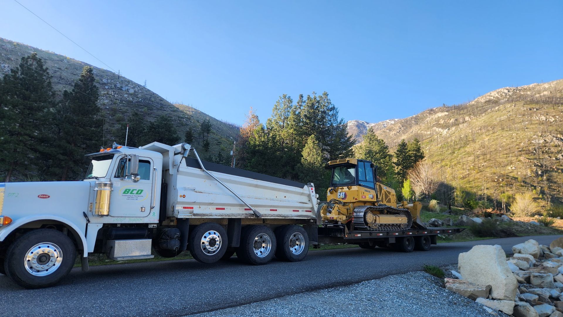 Tow truck hauling a yellow bulldozer on a flatbed beside a mountain road in daylight