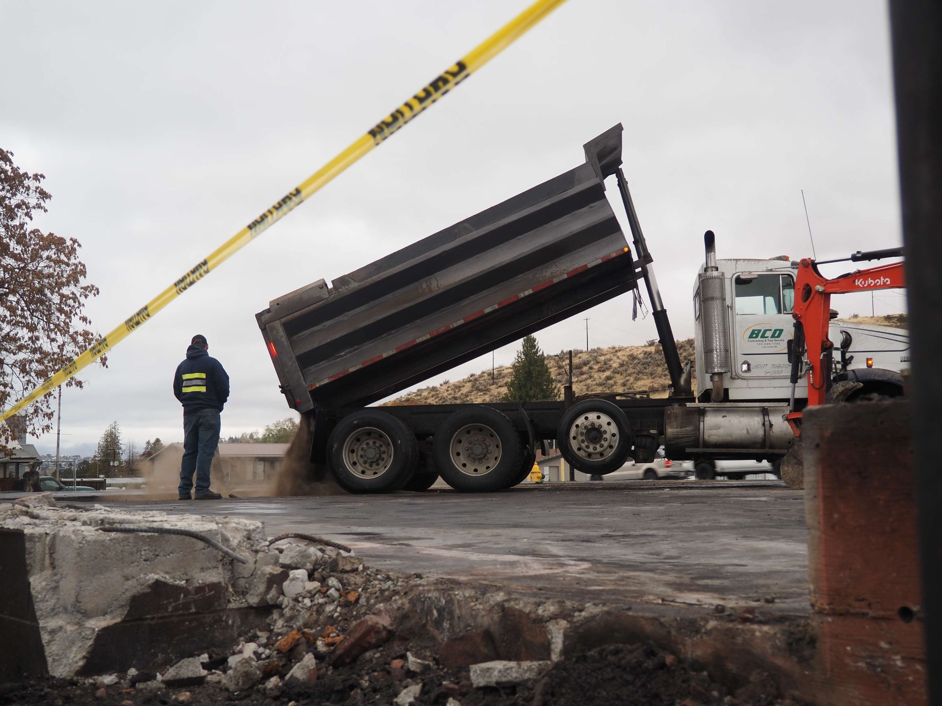 Dump truck raised at a roadside construction site, with a worker nearby and yellow caution tape overhead