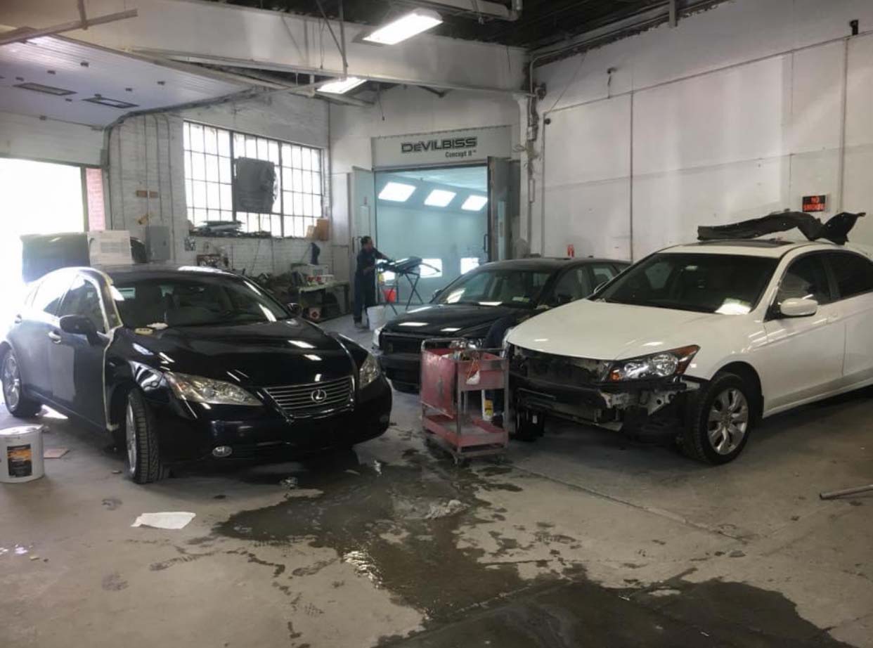 Two cars in a repair garage, a black sedan and a white SUV with front-end damage.