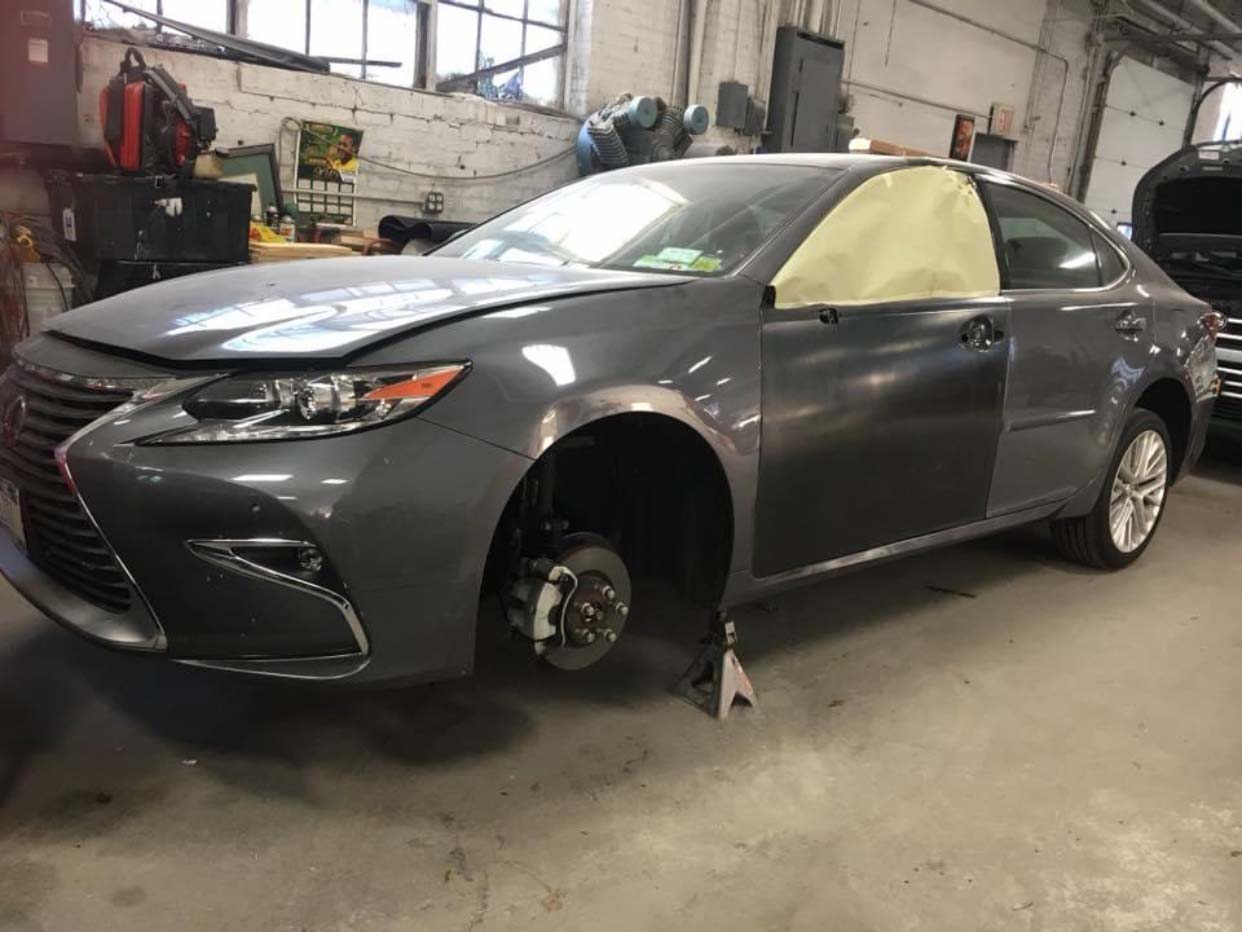 Gray sedan in a repair shop with the front wheel removed and the windows masked for painting