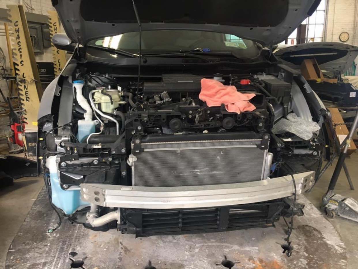 Front view of a heavily damaged car in a garage with its bumper removed and engine exposed