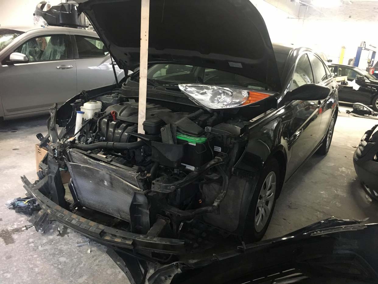 Severely damaged black car with hood open in a garage, front end crushed and torn off