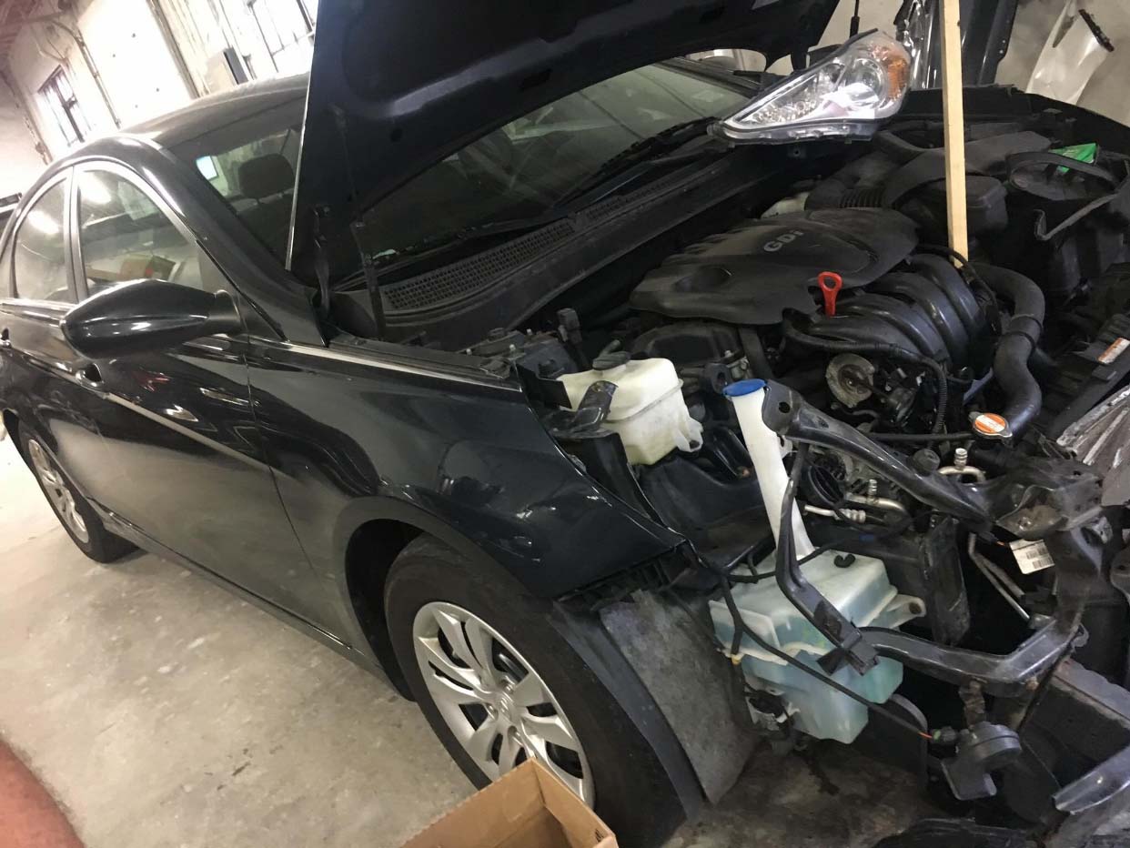 Black car with front-end crash damage in a garage, hood open and exposed engine visible