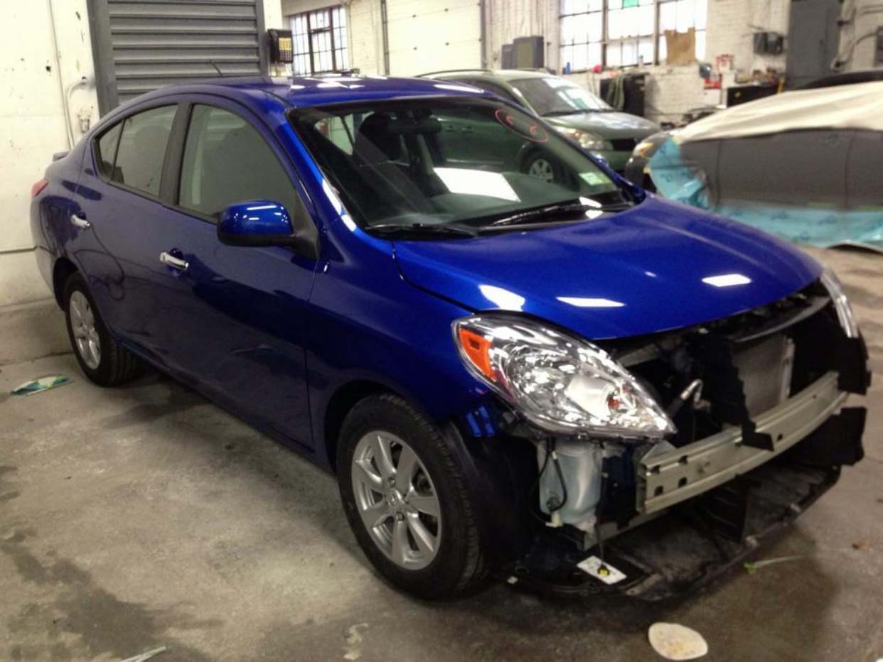 Blue sedan with damaged front bumper and missing grille in an indoor auto shop