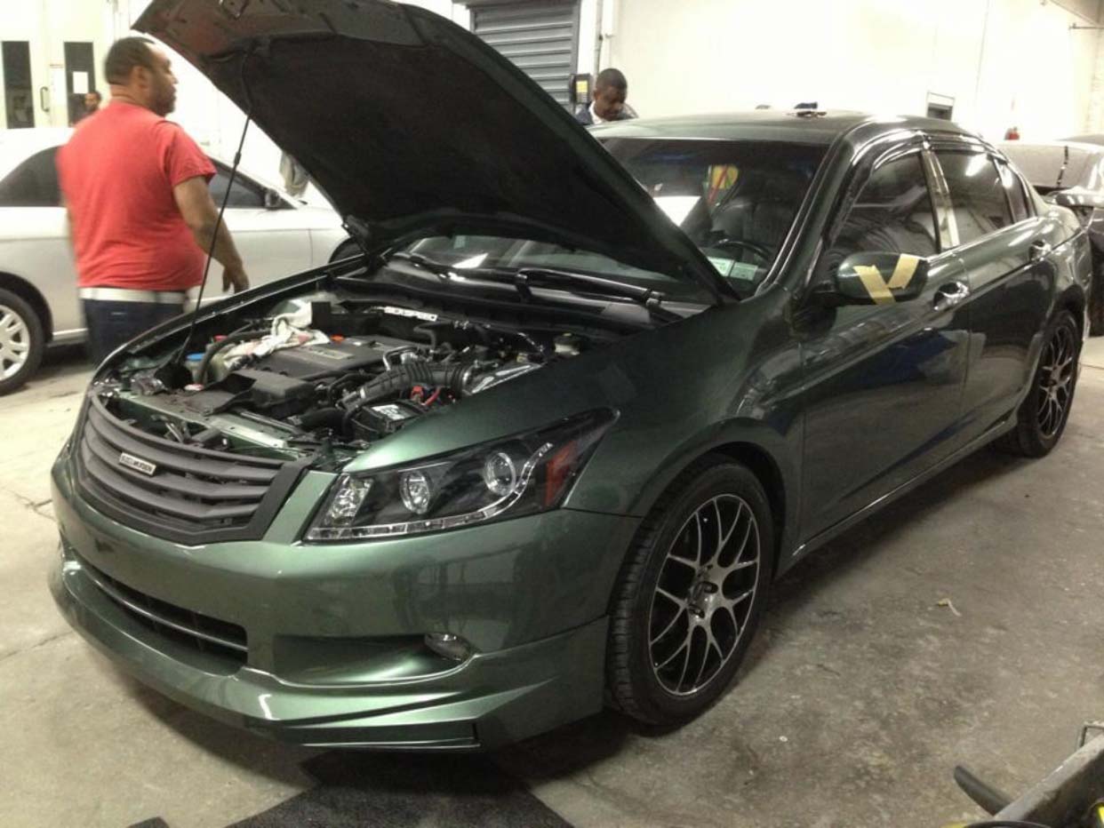 Dark green sedan with hood open in a garage, with a person standing beside it.