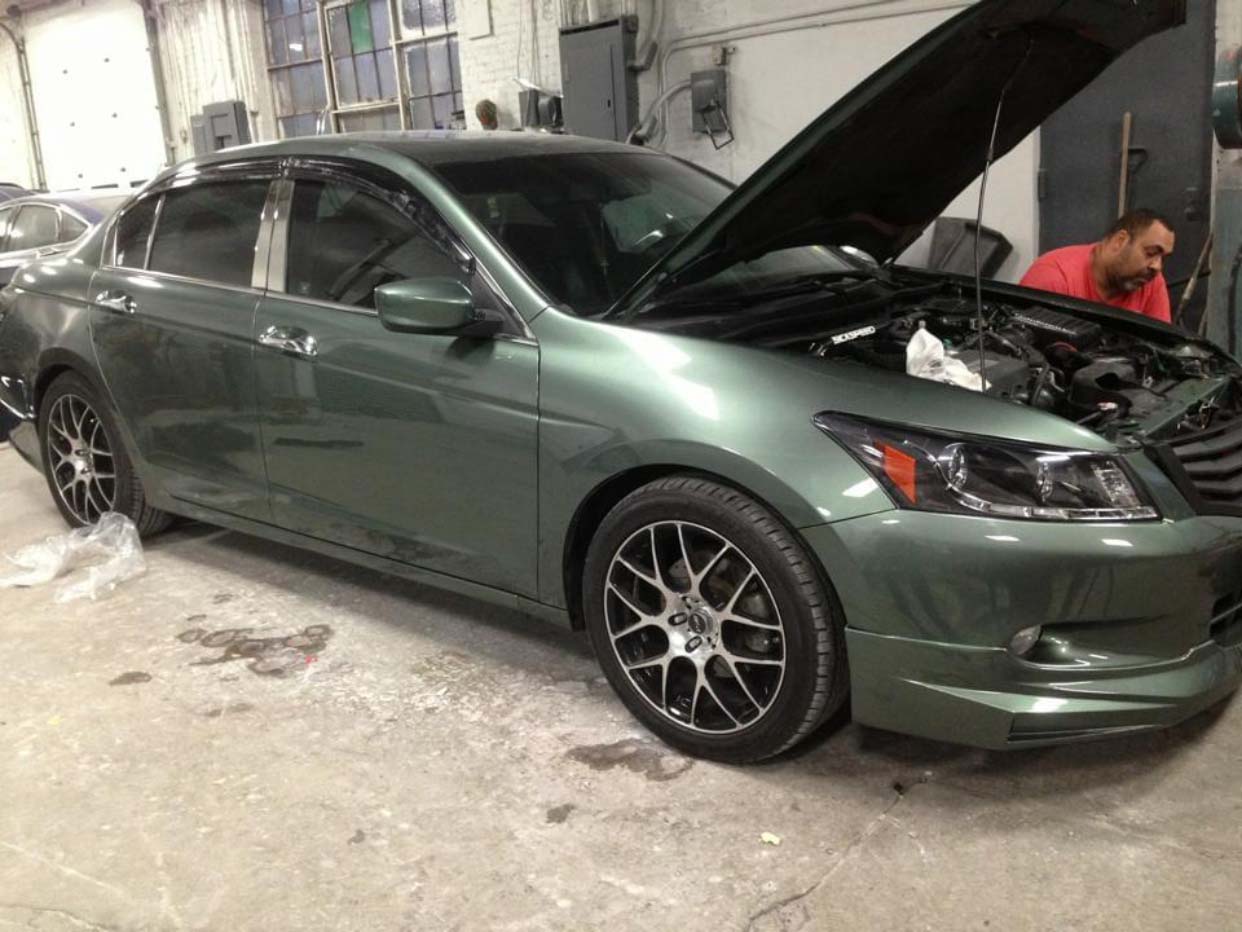 Green sedan with hood open in a garage, with a mechanic working beside it.