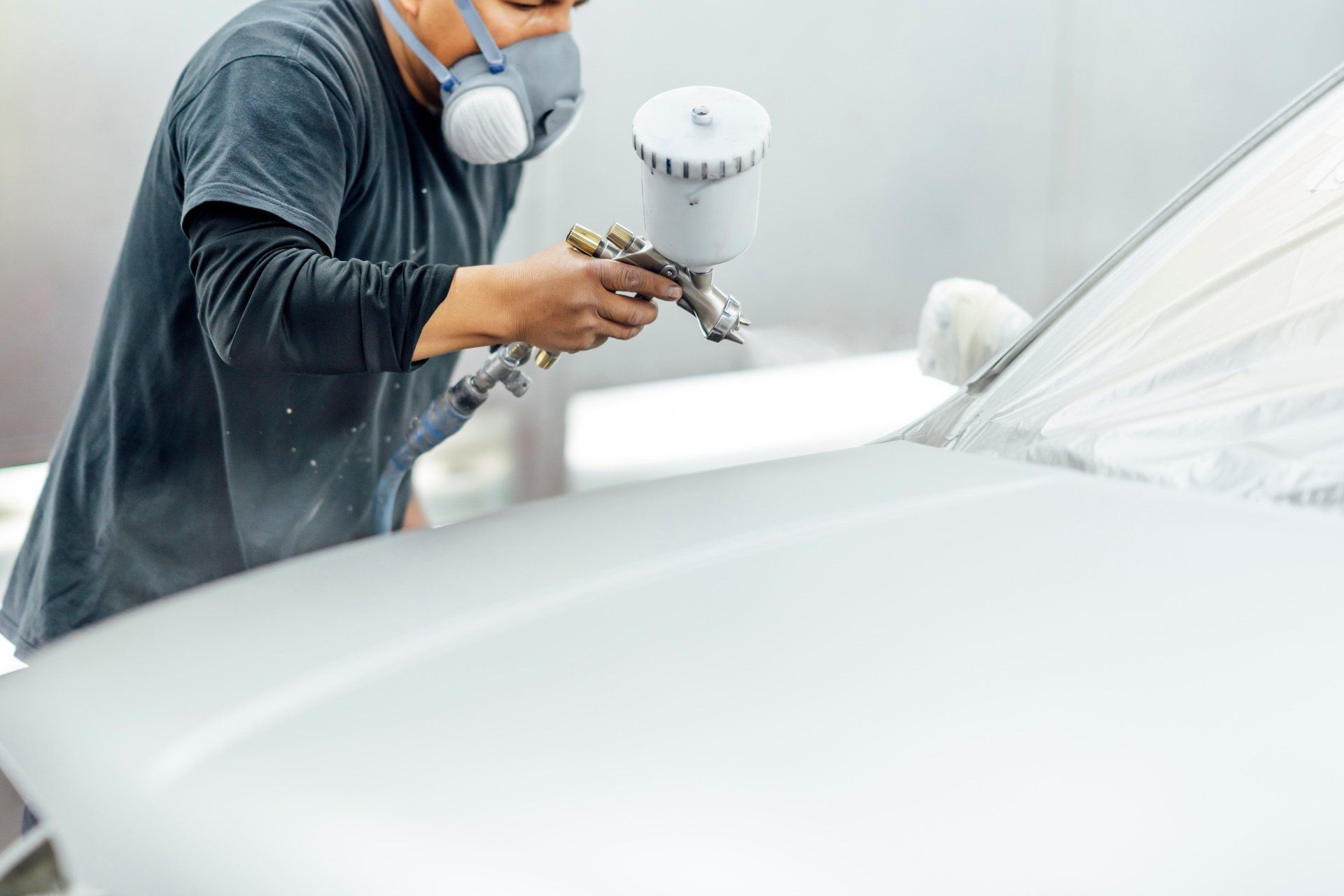 Worker spray-painting a white car in a paint booth, wearing a respirator mask