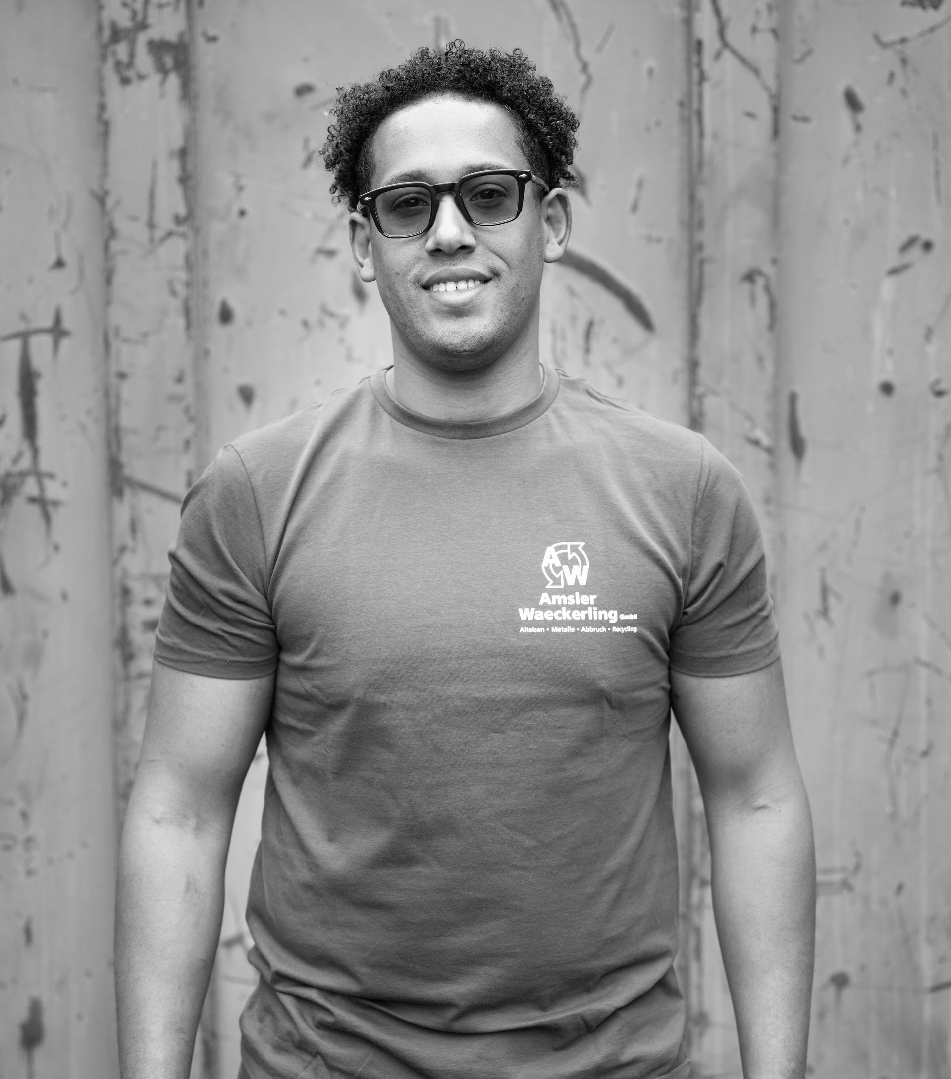 Ein Mann mit Brille und T-Shirt lächelt vor einer strukturierten Metallwand.