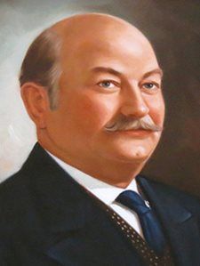 Portrait of a bald man with a mustache in a dark suit and blue tie, facing slightly right.