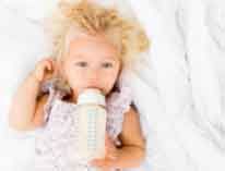 Blonde child lying on a white blanket, holding a baby bottle and looking at the camera.